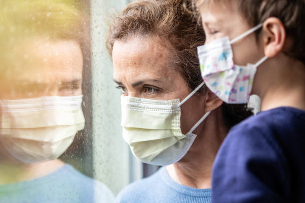 istock Mature woman posing with her son, both wearing protective masks, very sad looking through window worried about Covid-19 lockdown 1217022954