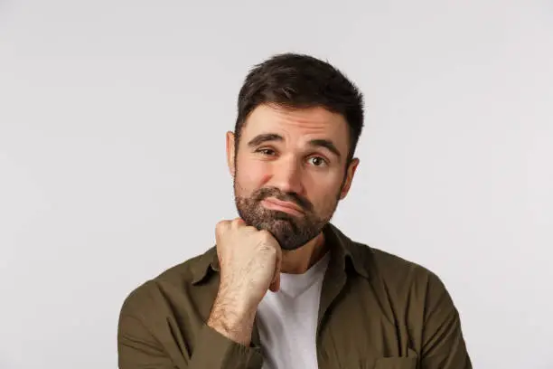 Bored and unimpressed, indifferent adult sad male with beard, looking reluctant and dissatisfied, lean head on fist, frowning, grimacing making indecisive upset expression, white background.