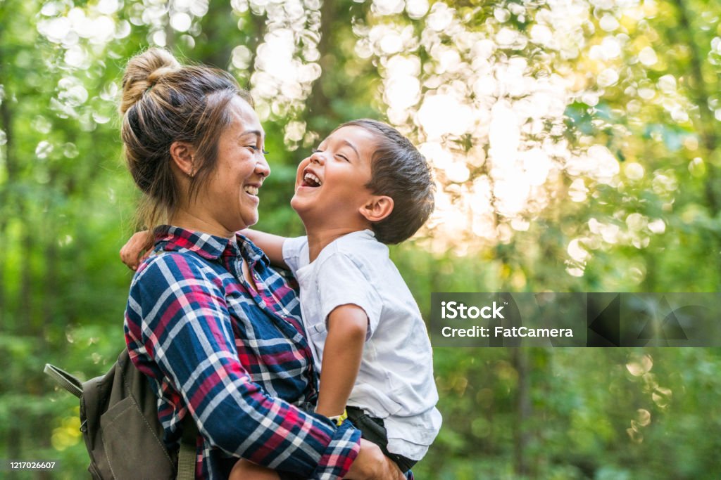 Mother And Son Enjoy Time in Nature stock photo A young mixed-race mother and son enjoy time in nature together.  The mother is holding her toddler son in her arms as they stand together in the woods.   The sun is shining through the green leaves of the trees and they are sharing a laugh together. Family Stock Photo