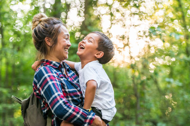 Mother And Son Enjoy Time in Nature stock photo A young mixed-race mother and son enjoy time in nature together. The mother is holding her toddler son in her arms as they stand together in the woods. The sun is shining through the green leaves of the trees and they are sharing a laugh together. indian man walking in park stock pictures, royalty-free photos & images