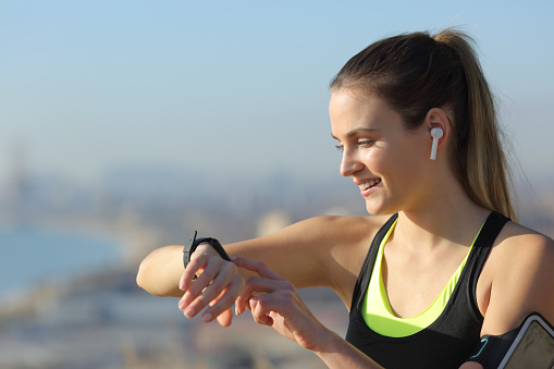 Runner wearing earbuds checks music on smartwatch outdoors in a city outskirts