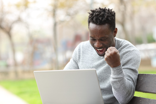 Excited black man celebrating checking laptop content sitting on a bench in a park