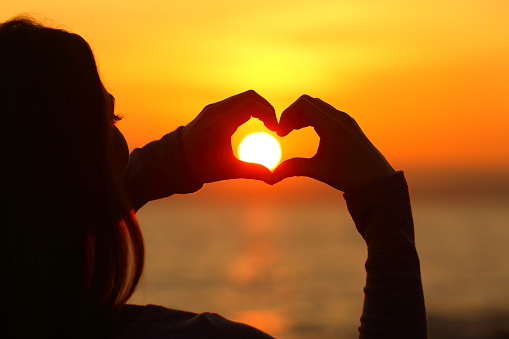 Close up of a girl hands silhouette making heart shape around the sun at sunset on the beach