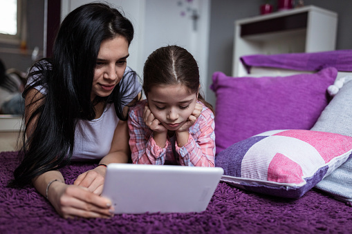 Teenage girl is watching with her younger sister a digital tablet while lying on the bedroom floor