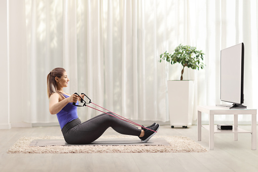 Woman sitting on the floor and exercising with a resistance band in front of a tv at home