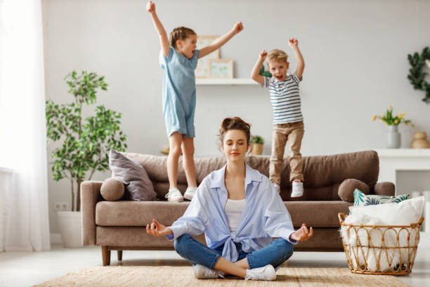 Tranquil young mother practicing yoga to stay calm with mischievous kids at home Happy mother with closed eyes meditating in lotus pose on floor trying to save inner harmony while excited children jumping on sofa and screaming in light spacious living room child behaving badly stock pictures, royalty-free photos & images