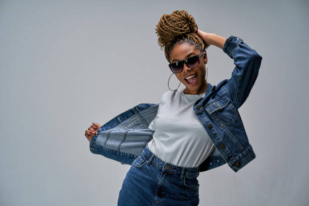 Happy girl in denim jacket sings and holds her hand on the back of her head. Emotions concept Woman with wide opened smile with bun of braids in earrings in sunglasses posing black woman hair bun stock pictures, royalty-free photos & images