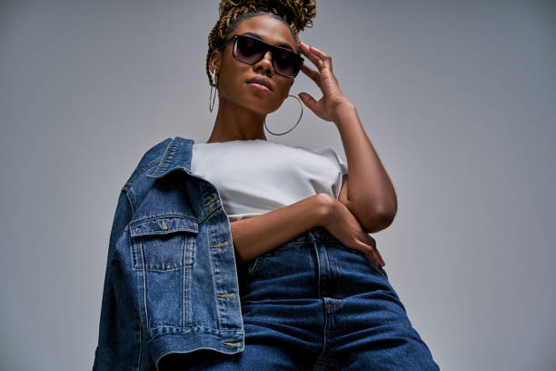 Confident lady in white t-shirt in jeans with denim jacket posing and looking at camera holds hands on glasses and hip. Fashion concept Woman in earrings with bun of braids holding a jacket on her shoulder in bottom view below photos stock pictures, royalty-free photos & images