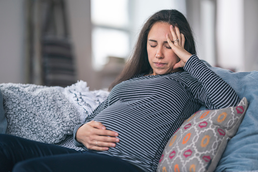 A pregnant Eurasian woman is in her second trimester. The woman is sitting on a couch and is holding her belly in discomfort. Prenatal, morning sickness, back pain, heartburn, constipation, and fatigue concepts.