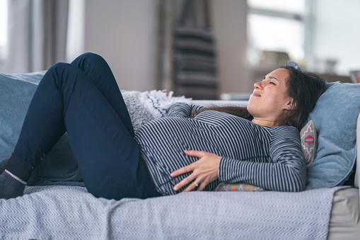 A mixed-race woman is pregnant. She is in her second trimester. The woman is lying on the couch in discomfort. She is holding her abdomen with both hands and has a pained expression on her face. Prenatal, morning sickness, back pain, heartburn, and constipation concepts.