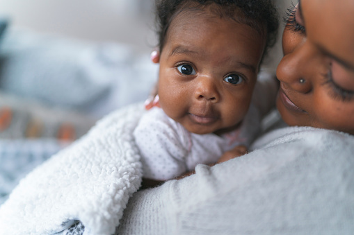 A young mother is cherishing special moments at home with her newborn daughter. The mother is tenderly holding her baby and looking at her lovingly. The baby is looking over her mother's shoulder directly at the camera. The close up image is focused on the content child. Family moments and postpartum concepts.