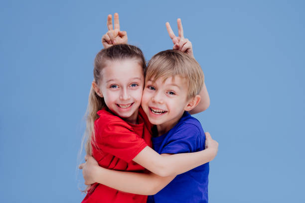 Cheerful cute little girl and boy in bright casual clothes hugging and making horns while looking at camera on blue background