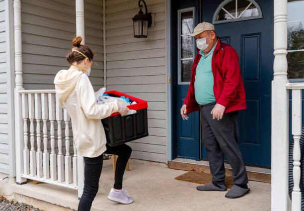 Young woman wearing protective n95 mask, a courier, is delivering the groceries packed into the reusable plastic bins to a customer, a senior man, during COVID-19 outbreak. Groceries home delivery during the COVID-19 coronavirus outbreak in Poconos, Pennsylvania, USA. home delivery photos stock pictures, royalty-free photos & images