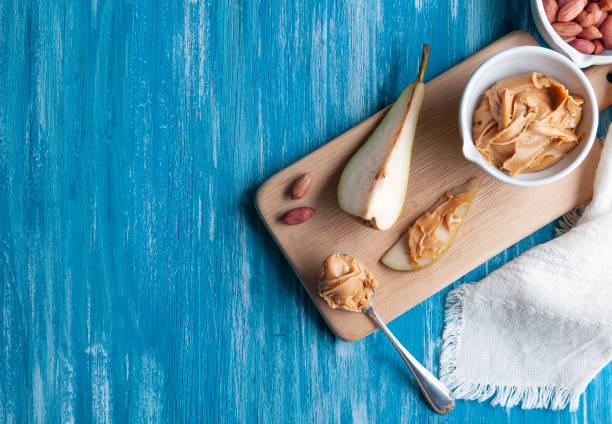 White cup with peanut butter. On a wooden board. Next to a chopped pear, peanuts and a spoon with pasta.
Flat lay composition. View from above. Blue wooden background. Space for text.
