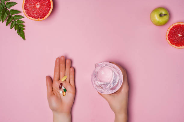 분홍색 배경에 여성의 손에 비타민과 물 한 잔. 텍스트에 배치합니다. 배경에 자몽, 사과와 녹색 잎. 건강한 라이프스타일 컨셉 - vitamin pill nutritional supplement capsule antioxidant 뉴스 사진 이미지