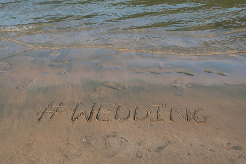 Image of hashtag tag writing on sunny beach in the soft sand, light by the early morning sunshine. Written using a stick by hand, with Wedding words as capital letters by the gentle lapping waves of the sea on the seashore. Picture taken at Palolem Beach, Goa, India, during the early morning golden hour without tourists on holiday vacation / beachgoers. Concept photo of Wedding handwriting in golden sand with modern hashtag prefix