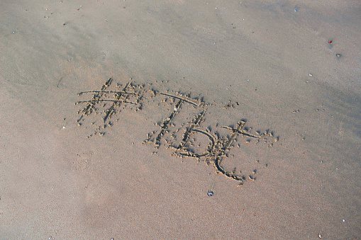 Image of hashtag tag writing on sunny beach in the soft sand, light by the early morning sunshine. Written using a stick by hand, with TBT stands for Throwback Thursday words as capital letters by the gentle lapping waves of the sea on the seashore. Picture taken at Palolem Beach, Goa, India, during the early morning golden hour without tourists on holiday vacation / beachgoers. Concept photo of TBT stands for Throwback Thursday handwriting in golden sand with modern hashtag prefix