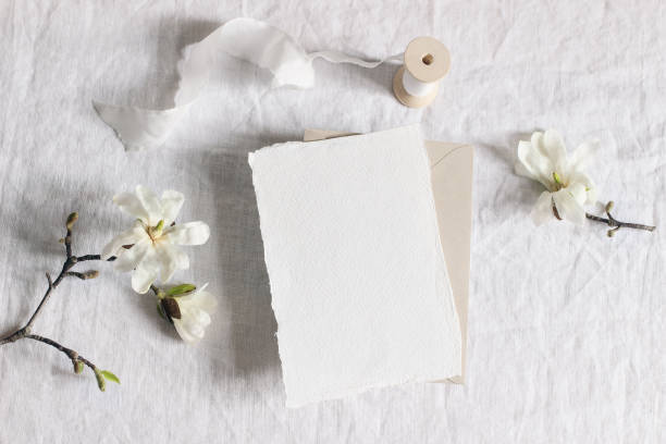 istock Wedding stationery mock-up scene. Blank vertical greeting card, envelope on linen tablecloth background with white magnolia stellata tree branches and ribbon. Feminine still life. Flat lay, top view. 1217110149