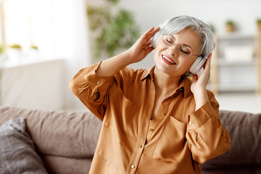 Elderly lady in headphones sitting on couch and listening to music with closed eyes while resting in cozy living room at home
