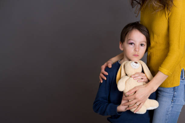 Sad boy with black eye, hurt bruised eye, hugging mom and teddy bear at home
