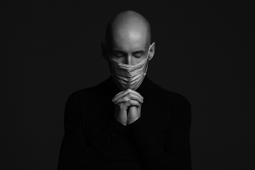 Coronavirus concept. Close up portrait of handsome bald man wearing medical mask, black turtleneck and praying. Studio shot