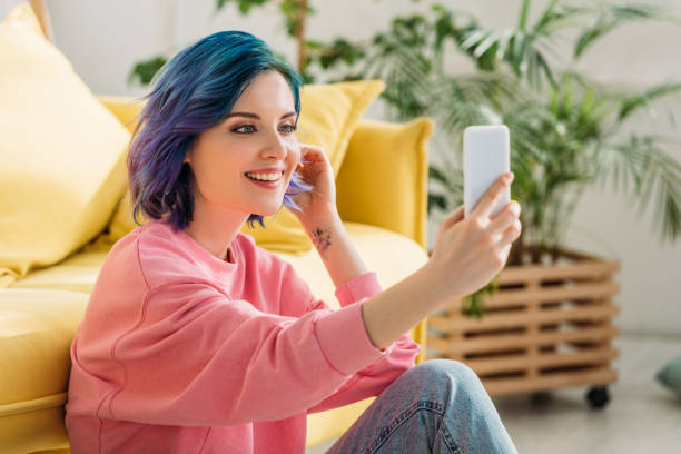 dziewczyna z kolorowymi włosami uśmiechnięta i robiąca selfie ze smartfonem w pobliżu sofy na podłodze w salonie - blue hair zdjęcia i obrazy z banku zdjęć