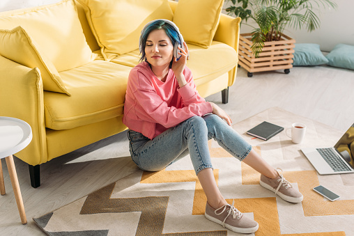 High angle view of freelancer with colorful hair, closed eyes and headphones smiling near sofa on floor