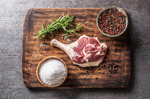 Top of view rustic wooden cutting board on dark background with duck thigh, herbs, spices and salt.