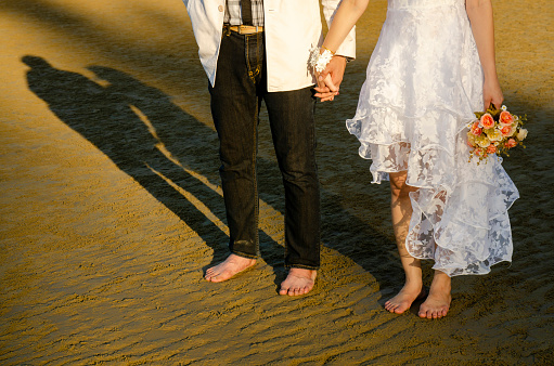 The bride and groom hold hands in the sandy desert beach at sunset, a beautiful wedding dress, long shadows, the wedding couple is enjoying each other, holding bridal bouquet