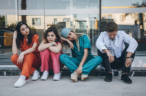 Stressed, tired or upset young doctor, nurse health care workers team with uniform looking unhappy sitting on a floor and taking break from work in behind of modern hospital building