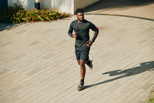 Serious young African-American sportsman running outdoors in shorts and rashguard when preparing for marathone