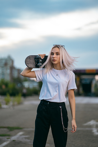 Teenage girl is standing outdoors with skateboard and looking at camera.