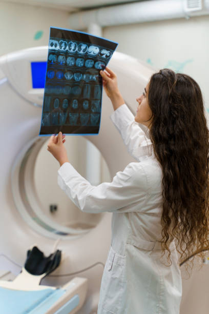 pionowy obraz studenta medycyny, który patrzy na zdjęcie rentgenowskie mózgu pacjenta w pobliżu kt komputer tomograhphy. młoda dziewczyna w klinice medycznej gotes nowy expirience - brain surgery mri scanner cat scan oncology zdjęcia i obrazy z banku zdjęć