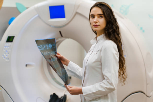 estudiante de medicina mira en cámara con imagen de rayos x del cerebro del paciente en sus manos. mujer se queda frente a kt komputer tomograhphy. joven en clínica médica tiene nueva experiencia - cat scan abdomen medical scan x ray fotografías e imágenes de stock