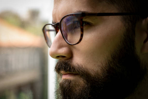 Quarantine self isolation concept. Young man staying at home, looking trough the window. Depressed man avoiding social contact. Worried person indoors activity. Anxiety. Agoraphobia.