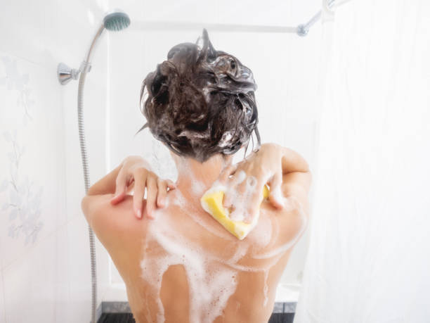 mujer desnuda con el pelo corto toma una ducha. la mujer se lava los hombros con una esponja amarilla. baño blanco. - shower gel fotografías e imágenes de stock