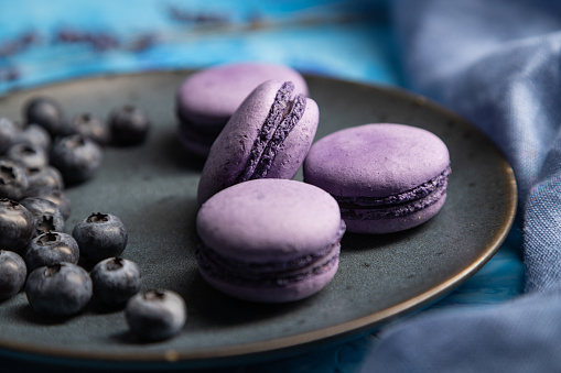 Purple macarons or macaroons cakes with blueberries on ceramic plate on a blue concrete background and blue textile. Side view, close up, selective focus.