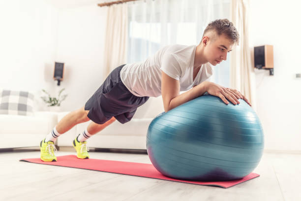 istock Working out at home by young man holding a plank position using a blue fit ball and red mat inside a living room. 1217169878