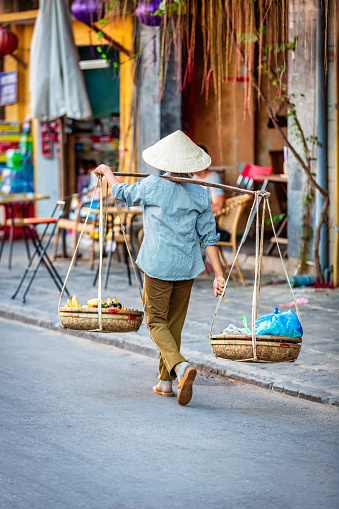 Hoi An, Vietnam - May 26th, 2019: Vietnamese Street Scene. Woman walking along the street in Hoi An carrying her street food baskets, walking by sidewalk cafe. Hoi An, Vietnam, East Asia