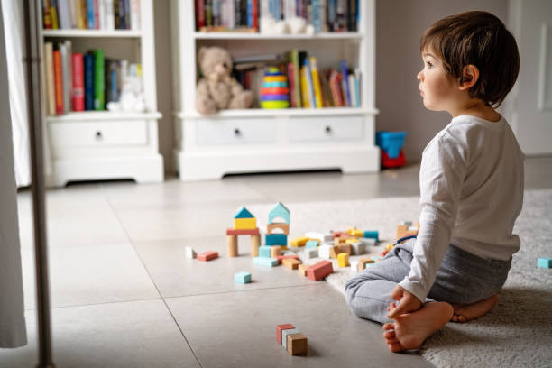 Little sad thoughtful bored toddler boy playing wooden colorful building blocks alone at home during quarantine looking at window. Child motor skills, creativity and imagination development game. Loneliness at self isolation period stock photo