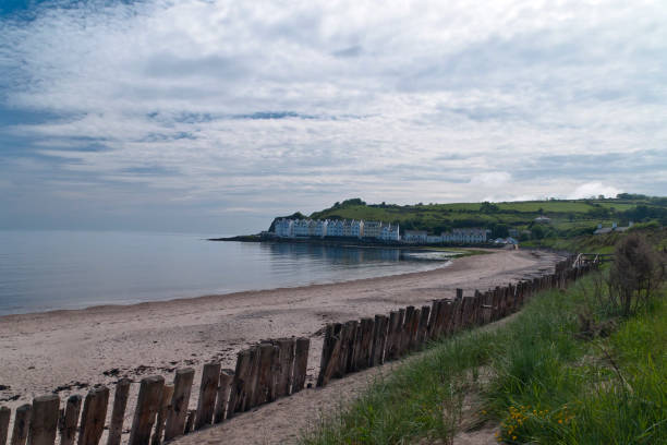 Cushendun Beach View South Cushendun (from Irish: Cois Abhann Doinne, meaning "foot of the River Dun") is a small coastal village in County Antrim, Northern Ireland. It sits off the A2 coast road between Cushendall and Ballycastle. It has a sheltered harbour and lies at the mouth of the River Dun and Glendun, one of the nine Glens of Antrim. Seen here is the harbor beach, a row of wood groynes, distant row houses. Cushendun is part of Causeway Coast and Glens district. cushendun michael stephen wills stock pictures, royalty-free photos & images