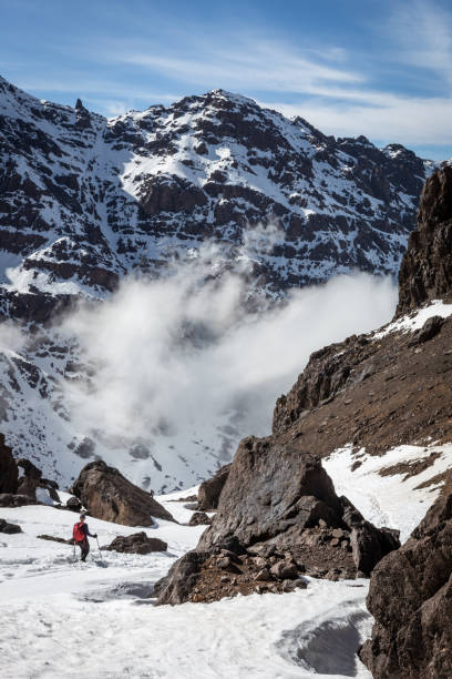 escursionista che scende nella neve in una valle sul monte toubkal, in marocco. in una giornata di sole con nuvole nella valle sottostante e paesaggio roccioso - atlas mountains foto e immagini stock