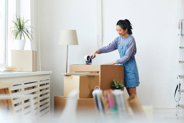Young Asian Woman Packing Boxes Side view portrait of young Asian woman packing cardboard boxes and smiling happily while moving to new home or apartment, copy space packing stock pictures, royalty-free photos & images