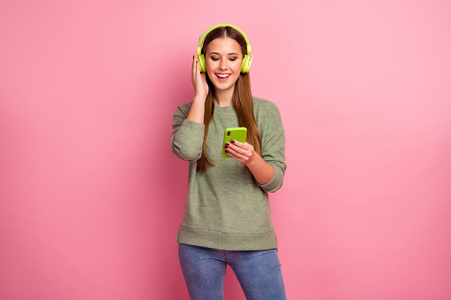 Portrait of excited energetic girl use smart phone, listen music on headset choose playlist enjoy wear sweater isolated over pastel color background