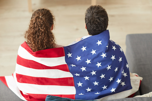 Back view portrait of young patriotic couple holding American flag while watching TV at home, copy space