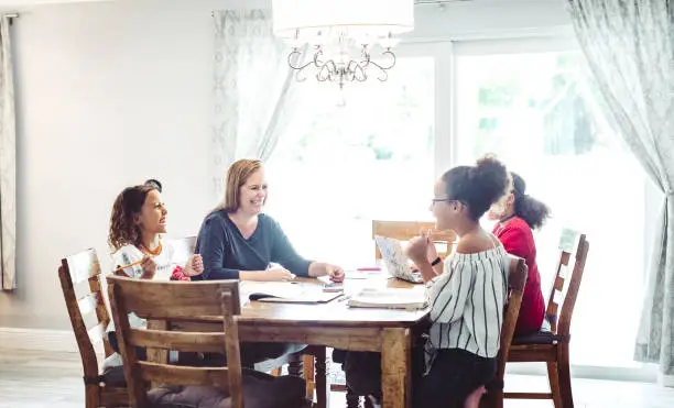 Blonde woman in her 40’s with three adolescent daughters sits at the family dining room table helping them with their schoolwork. They are doing homework, homeschooling or distance learning with mom’s help. Middle class happy mixed race family domestic life at home for a mom with her three children