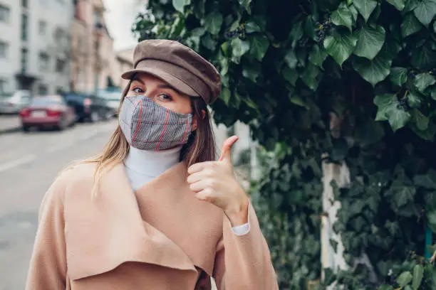Woman wears reusable mask outdoors during coronavirus covid-19 pandemic. Girl shows thumb up on empty street. Stay safe and positive. Spring fashion
