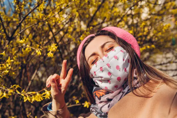 Woman wears reusable mask outdoors during coronavirus covid-19 pandemic. Girl taking selfie with spring flowers. Stay safe, positive. Spring fashion