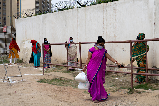Gurgaon, Delhi, India - circa 2020 : Underprivileged women in sarees stand in a ilne following the social distancing norms to collect food rations at a donation drive. The coronavirus pandemic has resulted in a lockdown of the country leaving people without a livelihood and depending on social handouts for survival