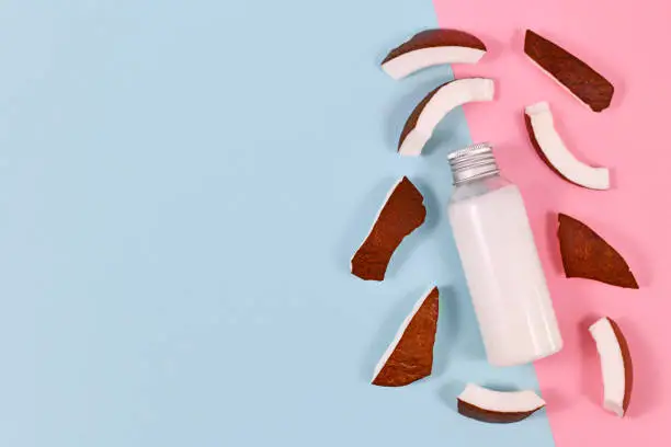 Photo of Bottle filled with coconut oil used for food preperation and cosmetic beauty products surrounded by slices of coconut fruit on blue and pink background